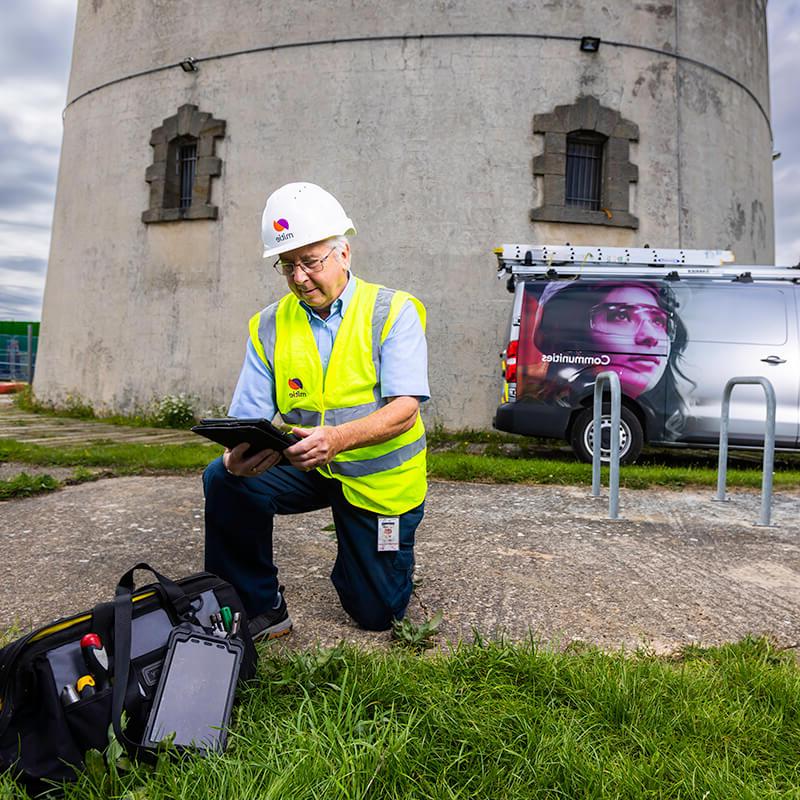 Mitie engineer kneeling on the ground next to a bag of tools, outside a tower in Essex County Council, while looking at an electronic tablet