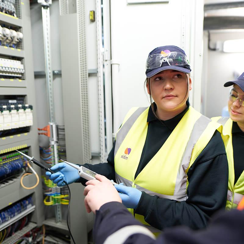 Two female engineer apprentices, wearing Mitie high vis vests and hats, in front of a wall of electrical wires and fuses