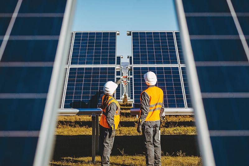 Two engineers in orange hi-ves vests and white hard hats stand in front of a field of solar panels